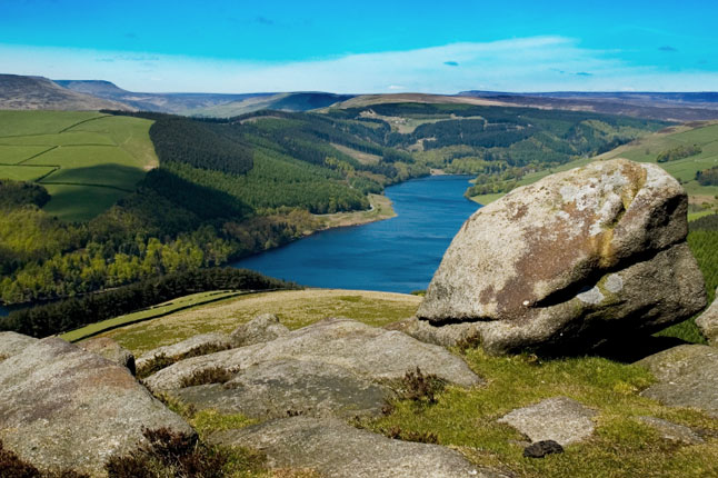 Panoramic vista of a calm lake and rugged mountains, framed by the gentle slope of a hill in the foreground