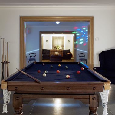 A pool table with balls arranged for a game sits in a room, with pool cues on a stand to the left. In the background, a cosy party house vibe emerges from sofas and colorful lights visible through a large doorway.