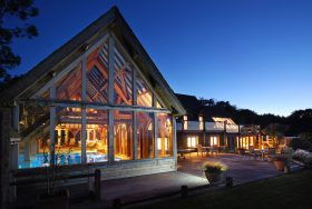 Modern house with large glass windows, warmly lit interiors, and an outdoor deck with seating, set against a twilight sky