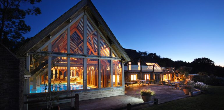 Modern house with large glass windows, warmly lit interiors, and an outdoor deck with seating, set against a twilight sky