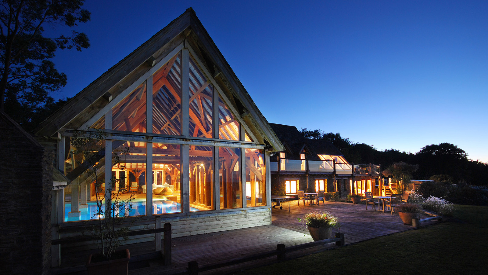 Modern house with large glass windows, warmly lit interiors, and an outdoor deck with seating, set against a twilight sky