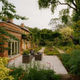 A serene garden featuring a patio adjacent to a charming brick building, surrounded by lush greenery and vibrant flowers